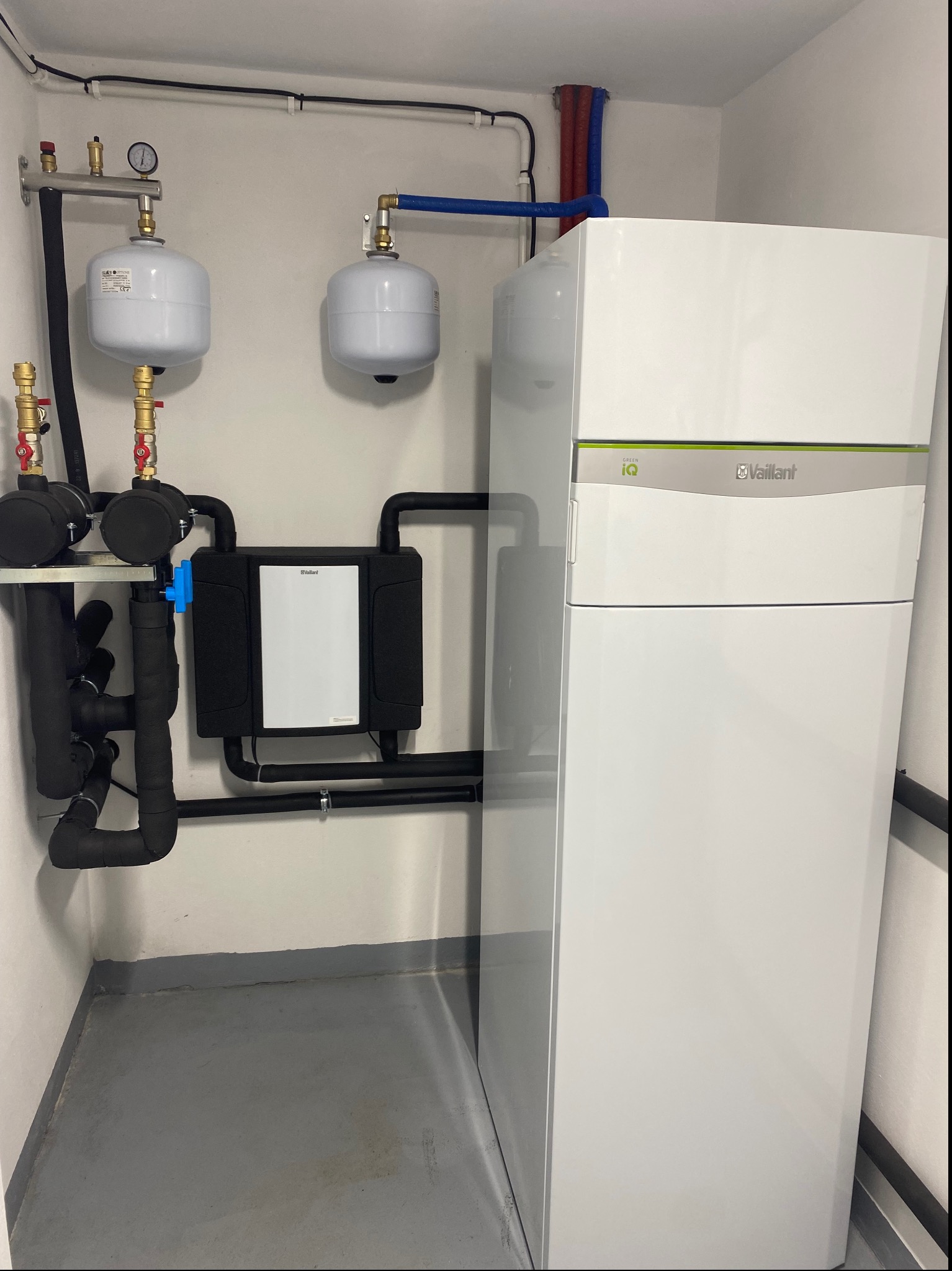 Biała pompa ciepła Vaillant Green iQ z podłączonymi rurami, zbiornikami wyrównawczymi i osprzętem hydraulicznym na tle białej ściany i szarej podłogi w kotłowni.