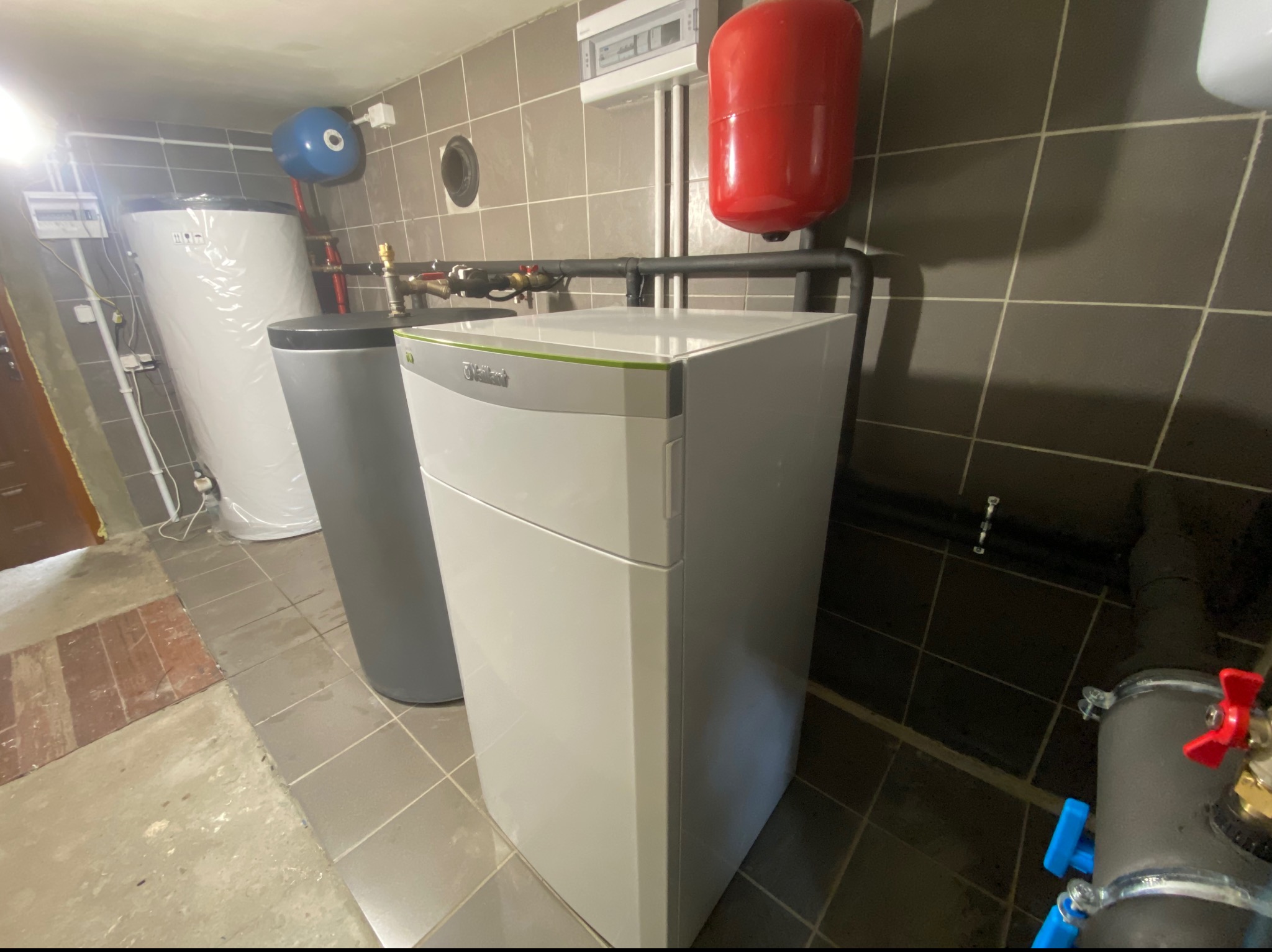 Biały piec grzewczy Vaillant z szarym akcentem, obok szary zbiornik buforowy i biały zasobnik ciepłej wody. Czerwony zbiornik wyrównawczy na ścianie z szarymi płytkami. Widoczne rury instalacji...