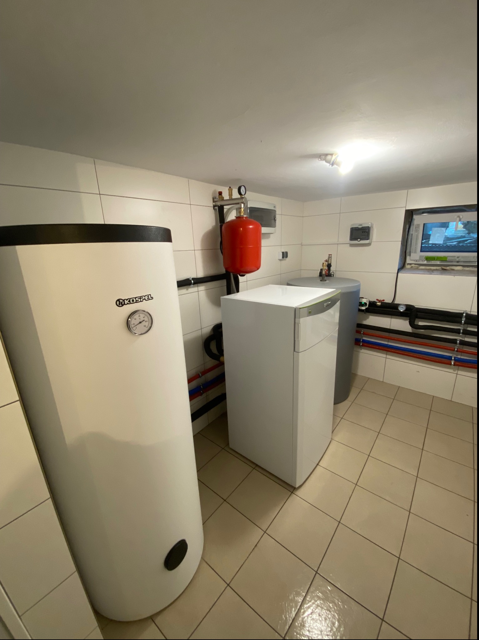 Kotłownia z białym zasobnikiem ciepłej wody Kospel z manometrem, szarym zbiornikiem wyrównawczym, czerwonym naczyniem przeponowym i instalacją z rur w kolorach niebieskim i czerwonym na tle białych...