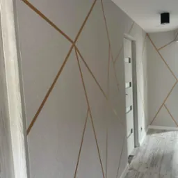 Ściana w korytarzu z dekoracyjnym malowaniem w geometryczne wzory w odcieniach bieli i złota, drzwi wewnętrzne z przeszkleniami i podłoga imitująca drewno.