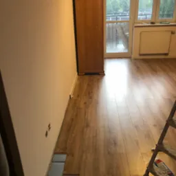 Fragment pokoju w trakcie montażu podłogi z paneli, widoczna warstwa izolacyjna, ściana z gniazdkami elektrycznymi oraz drabina oparta o podłogę.