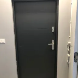 Szare drzwi z trzema poziomymi panelami, srebrną klamką i zamkiem, widoczne przed malowaniem ściany na szaro, z widocznymi kablami elektrycznymi i włącznikiem światła.