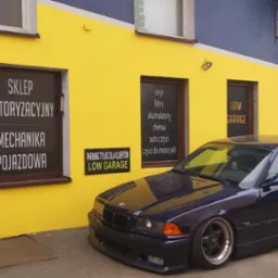 Niebieskie BMW E36 coupe zaparkowane przed warsztatem samochodowym 'LOW GARAGE' z żółto-niebieską fasadą, widoczne szyldy z ofertą sklepu motoryzacyjnego i usług warsztatowych.