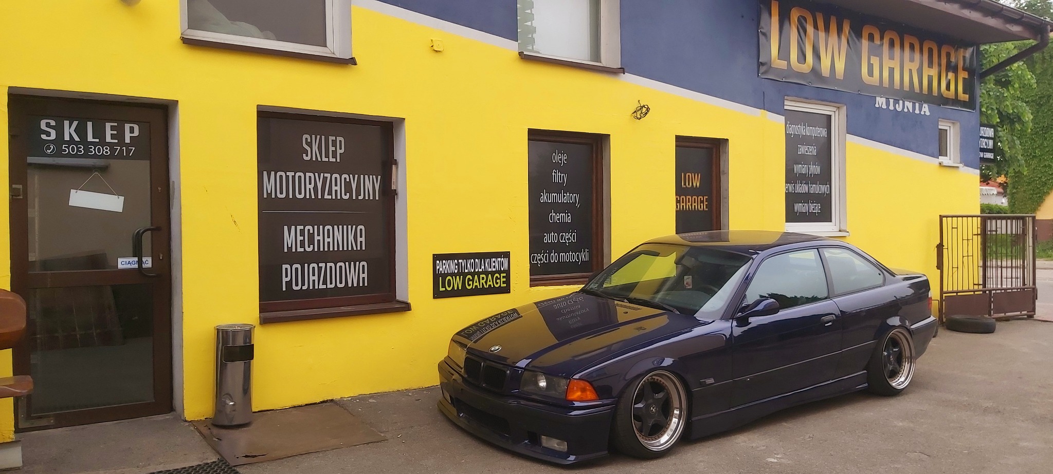 Niebieskie BMW E36 coupe zaparkowane przed warsztatem samochodowym 'LOW GARAGE' z żółto-niebieską fasadą, widoczne szyldy z ofertą sklepu motoryzacyjnego i usług warsztatowych.