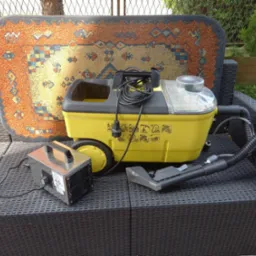 Żółty odkurzacz piorący z wężem i końcówką oraz generator ozonu na czarnym rattanowym krześle ogrodowym, za nimi dywan z wzorem etnicznym i fragment zieleni.