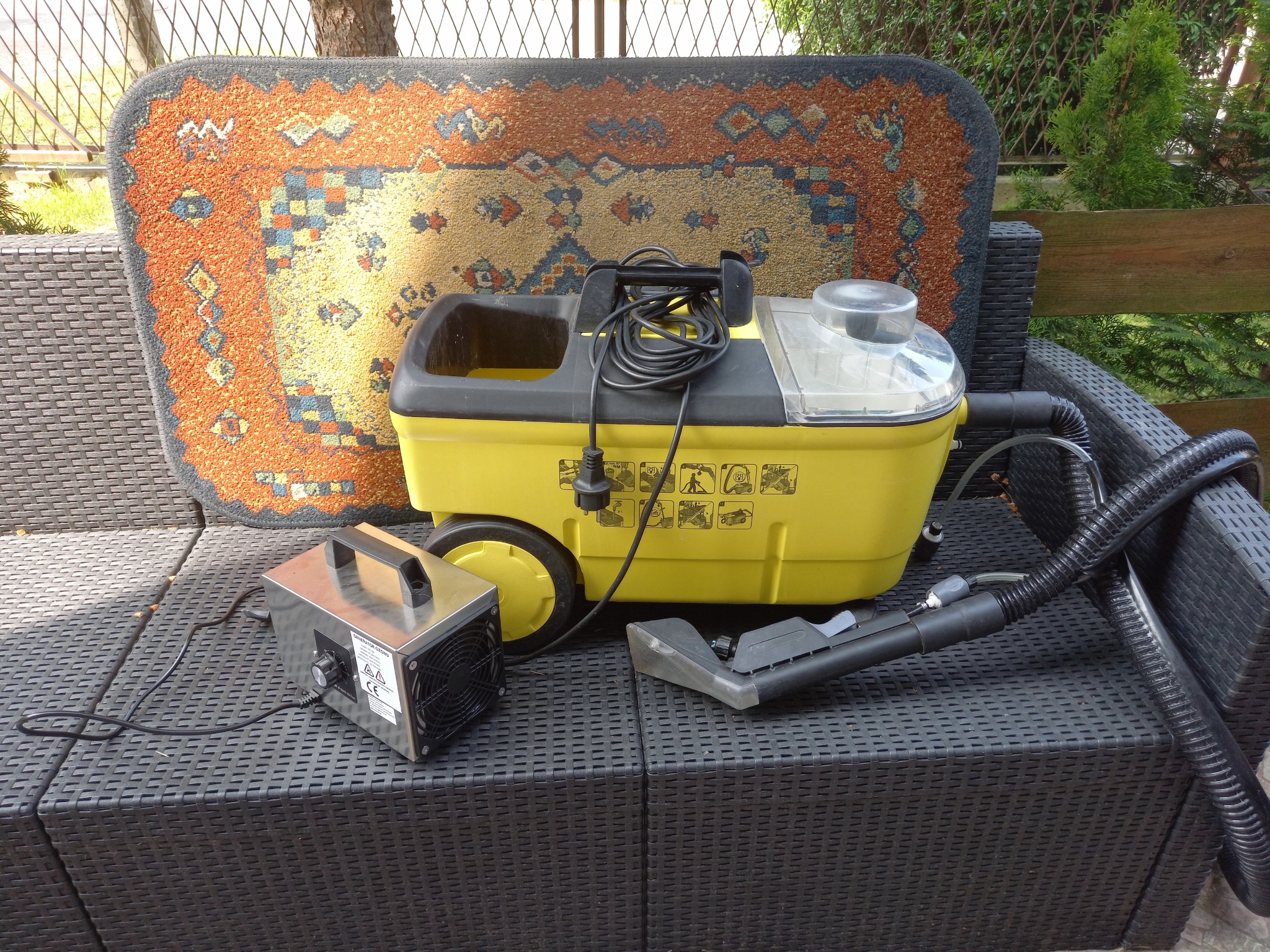 Żółty odkurzacz piorący z wężem i końcówką oraz generator ozonu na czarnym rattanowym krześle ogrodowym, za nimi dywan z wzorem etnicznym i fragment zieleni.