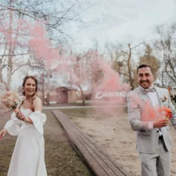 Uśmiechnięta para młoda w plenerze z kolorowym dymem: panna młoda w białej sukni z bukietem, pan młody w szarym garniturze z muszką, różowy dym w tle drzew.