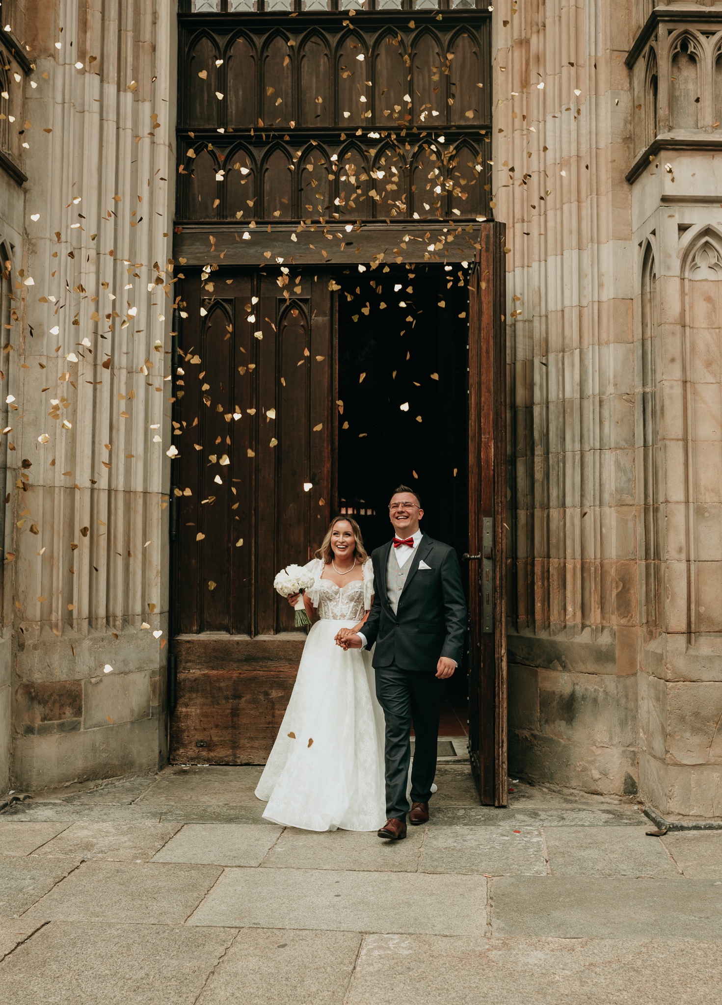 Para młoda wychodzi z kościoła obsypana złotym konfetti w kształcie serc, panna młoda trzyma bukiet białych róż.