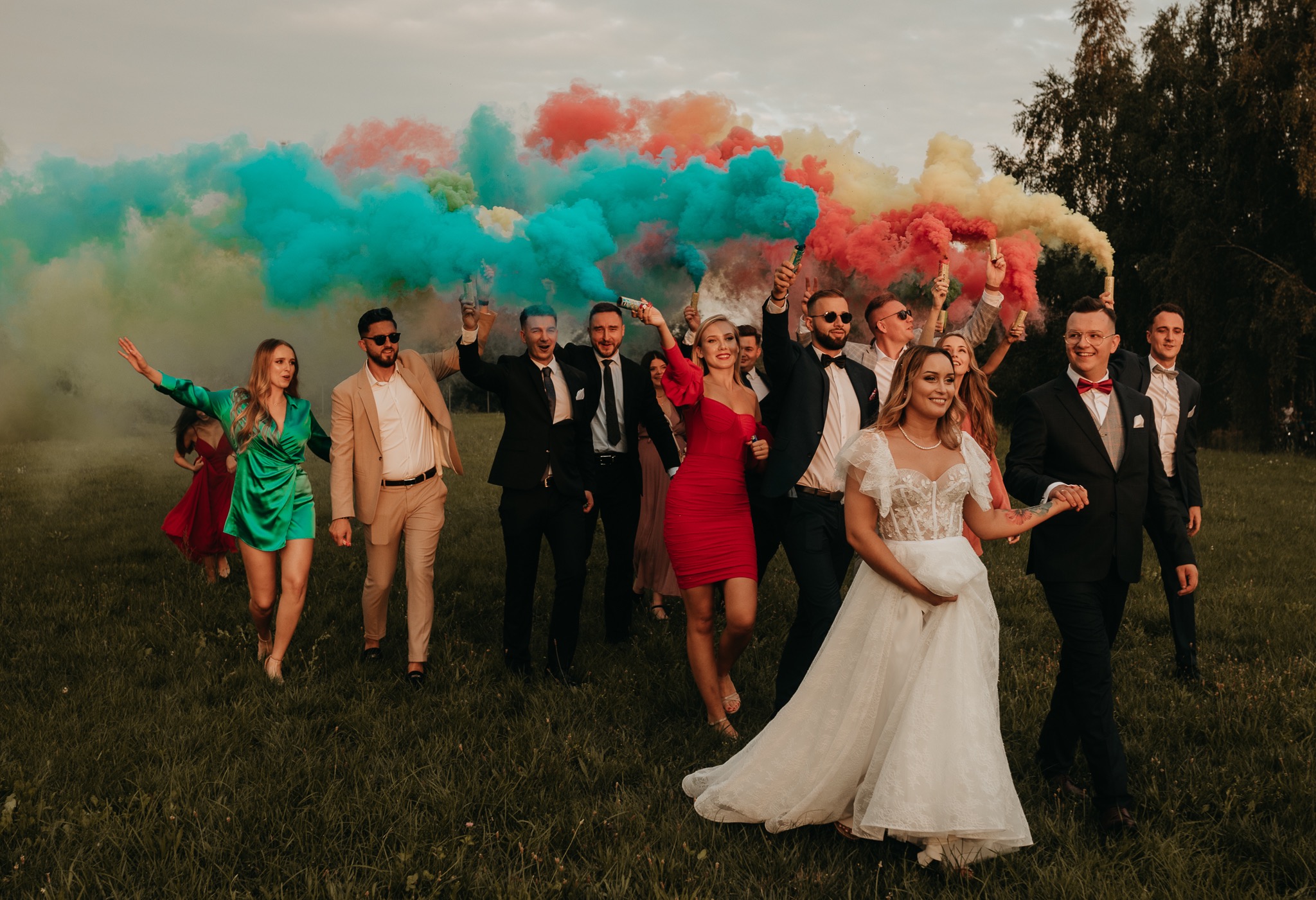 Grupa weselników z kolorowymi dymami na łące; panna młoda w koronkowej sukni, pan młody w smokingu, goście w strojach wieczorowych, w tle drzewa i pochmurne niebo.