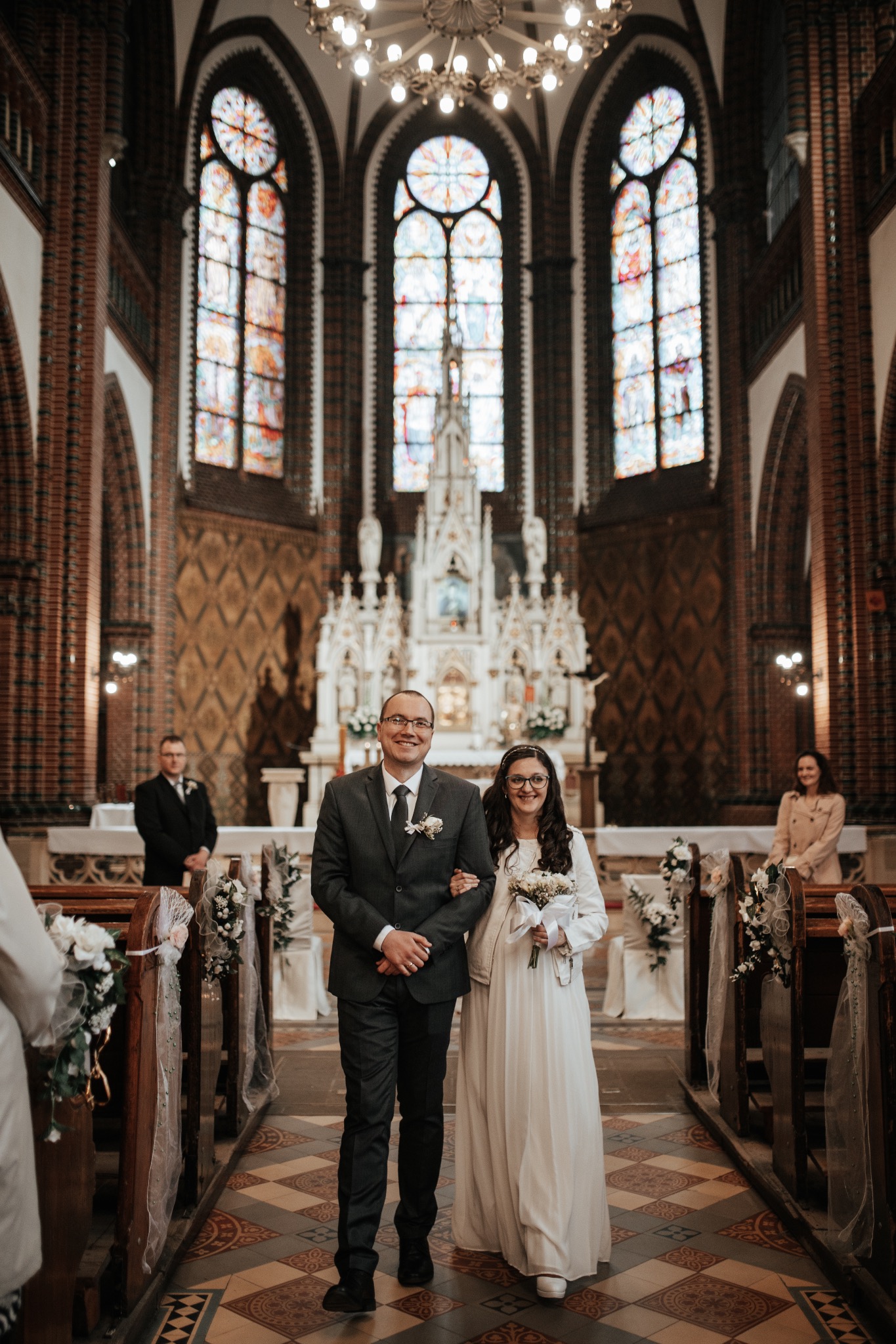 Para młoda, mężczyzna w szarym garniturze i kobieta w białej sukni, idą nawą kościoła w kierunku ołtarza, witraże w tle.