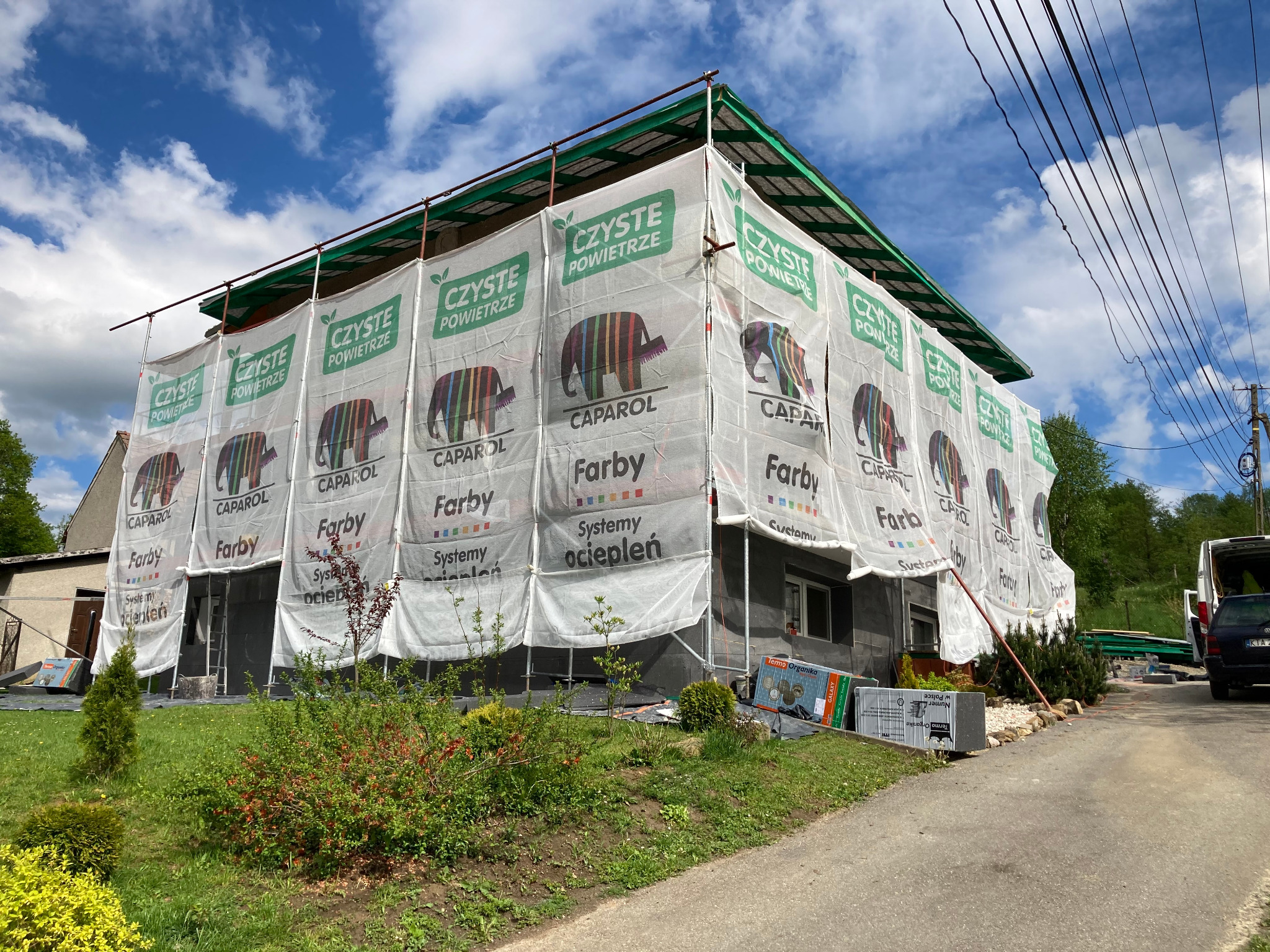 Dom jednorodzinny w trakcie ocieplania, osłonięty siatką z logo producenta farb, widoczne rusztowania i materiały budowlane na posesji.