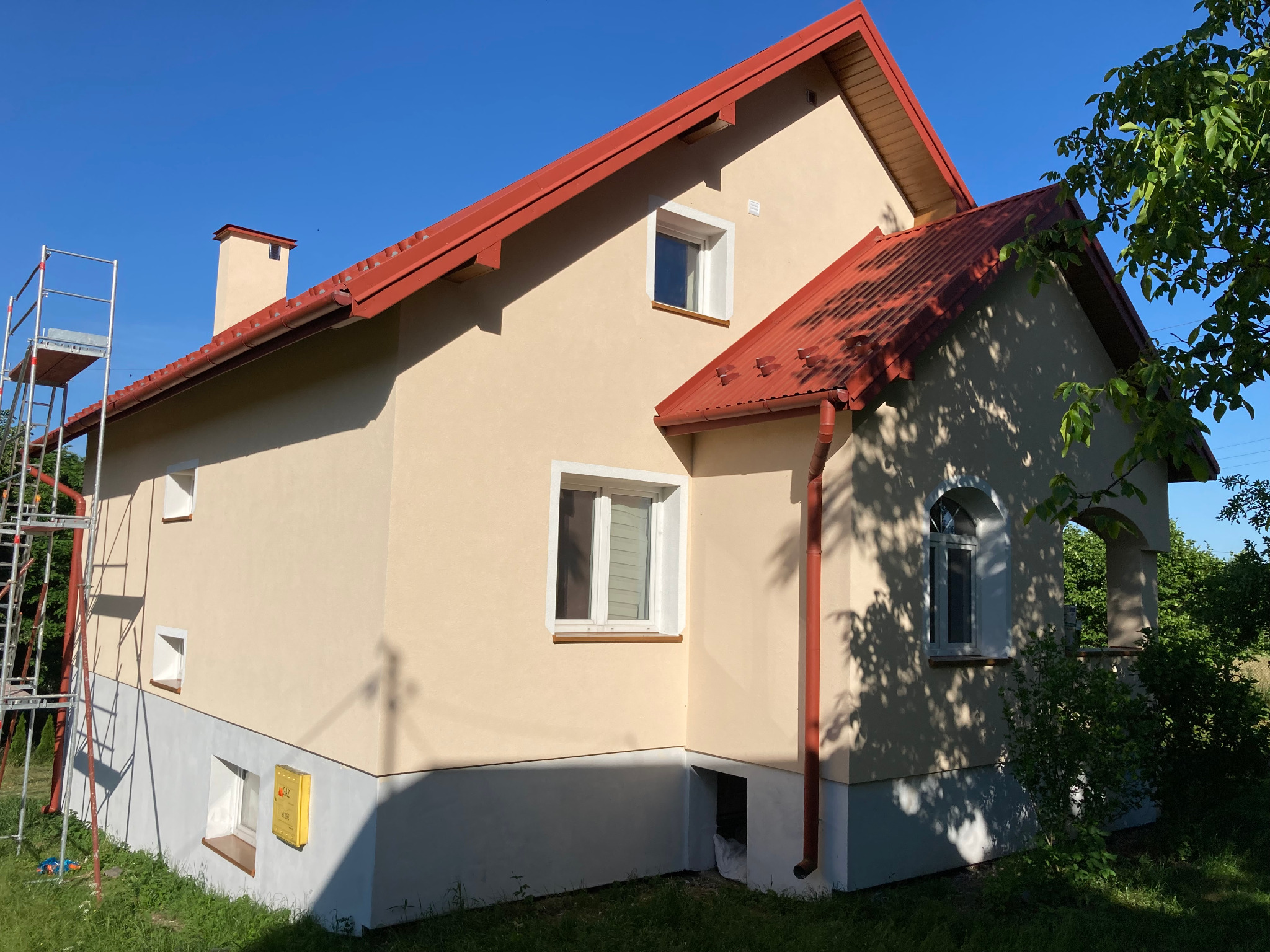 Dom jednorodzinny z beżową elewacją i czerwoną blachodachówką, widoczny fragment rusztowania przy ścianie bocznej, żółta skrzynka gazowa na elewacji, okna z białymi ramami.