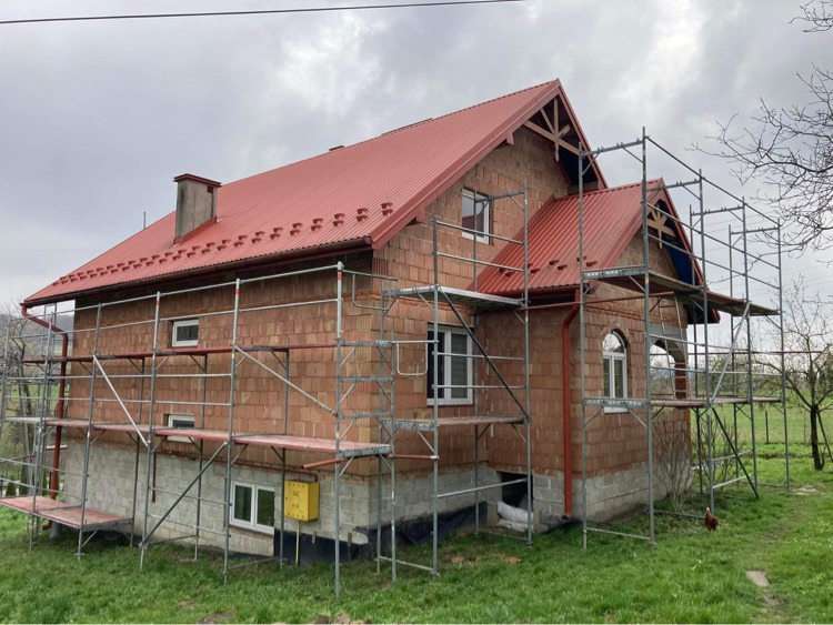 Dom w trakcie budowy, z czerwonym dachem i rusztowaniem, widok z boku na elewację z pustaków.