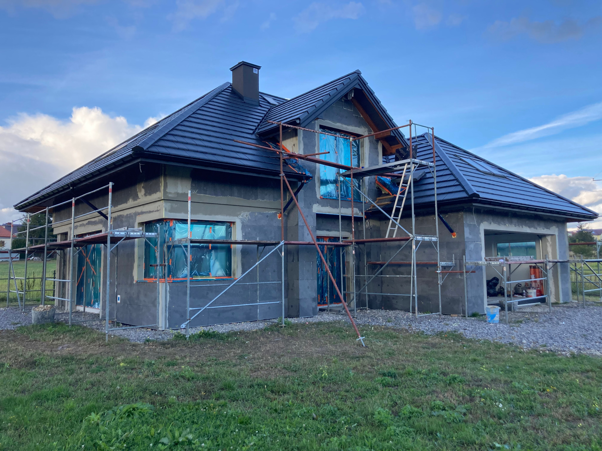 Dom jednorodzinny w trakcie ocieplania, zabezpieczone okna folią, rusztowania wokół budynku, widoczne płyty izolacyjne na ścianach.