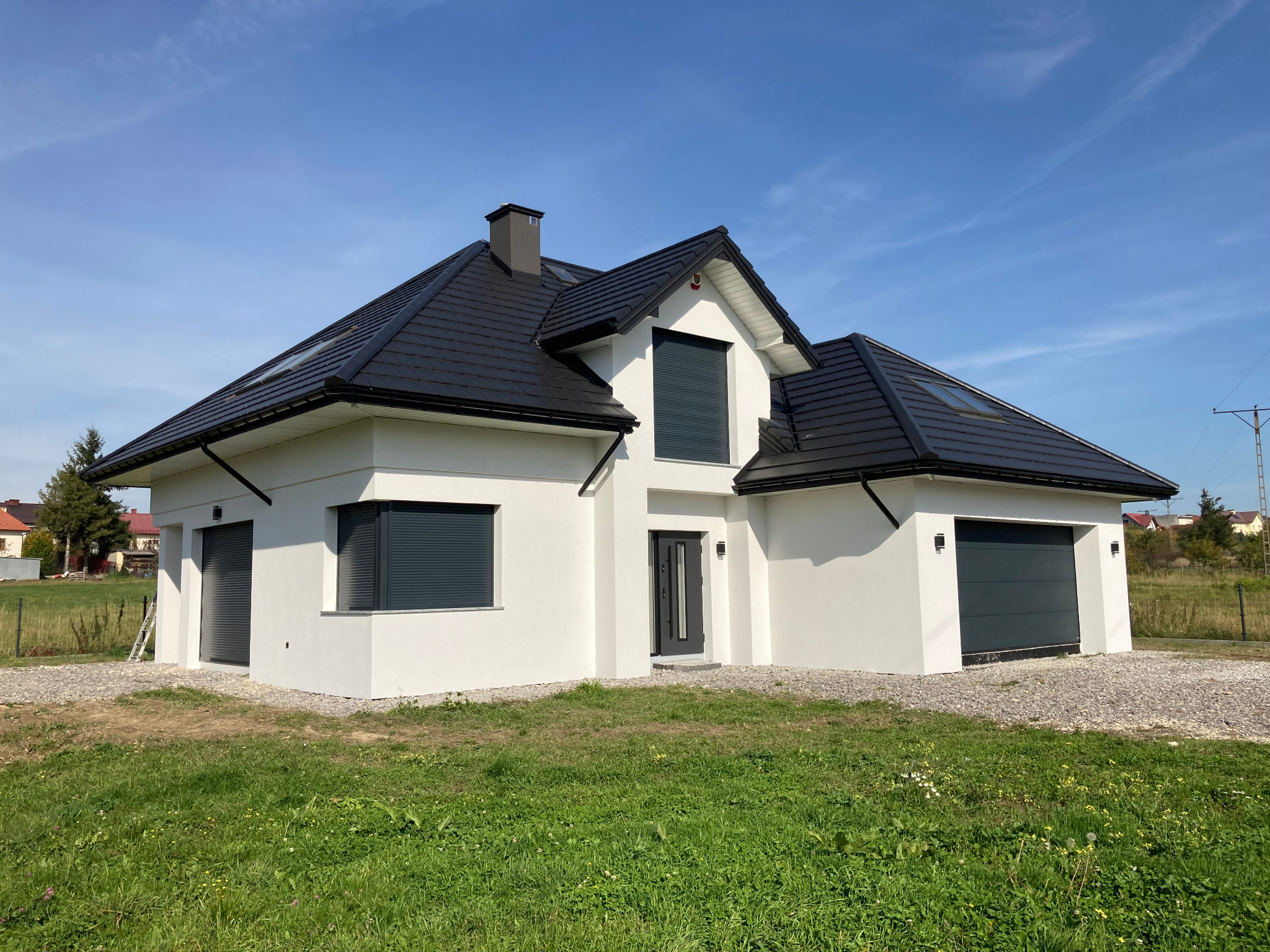 Nowoczesny dom jednorodzinny z białą elewacją, grafitowym dachem i roletami zewnętrznymi, otoczony trawnikiem i żwirowym podłożem.