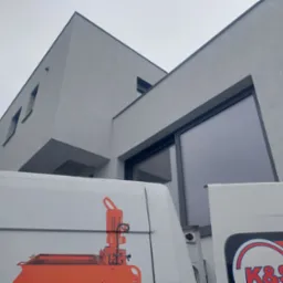 Nowoczesny, szary dom jednorodzinny z dużymi oknami, świeżo po nałożeniu tynku zewnętrznego, widoczny fragment furgonetki z logo firmy specjalizującej się w tynkach.