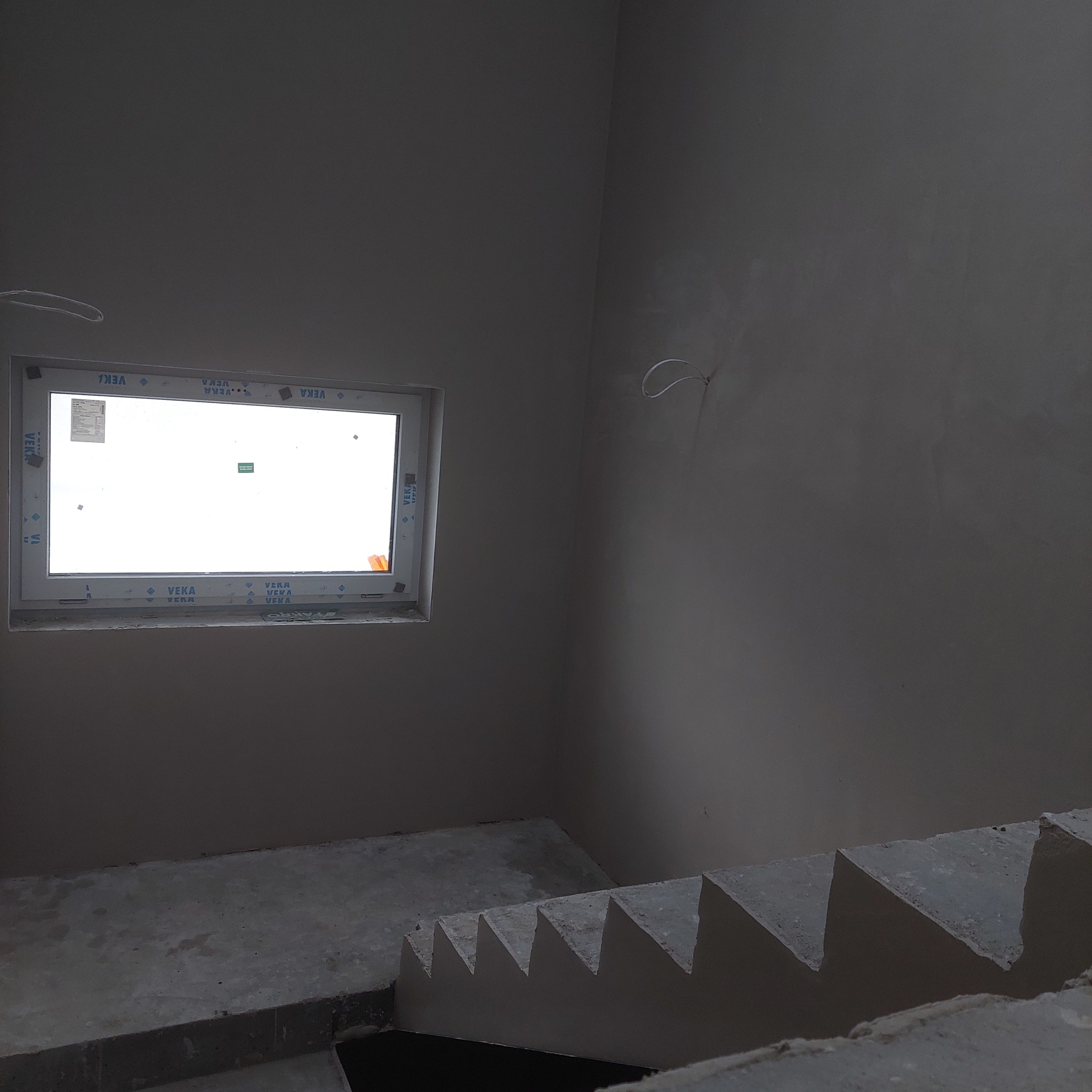 Surowe, szare wnętrze w trakcie prac wykończeniowych: świeżo otynkowana ściana z oknem Veka i betonowe schody w stanie surowym.