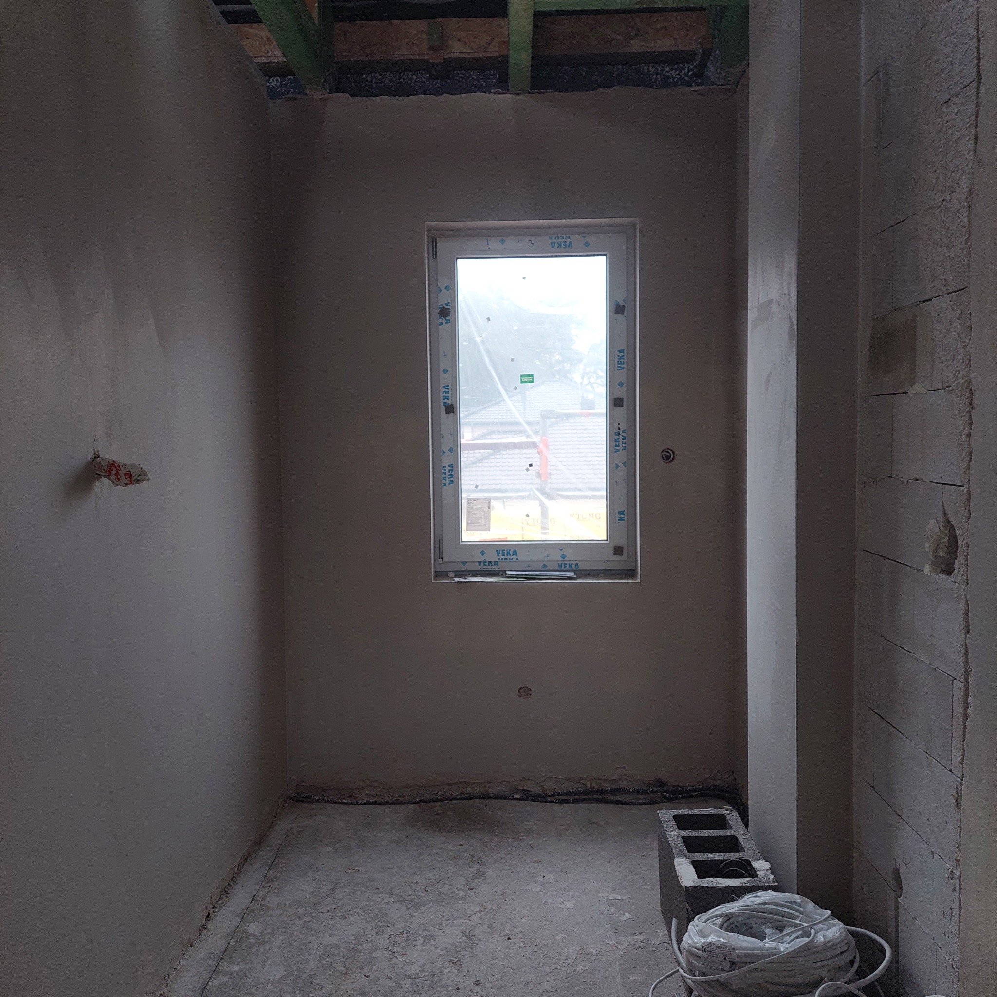 Wnętrze w trakcie prac tynkarskich: ściany pokryte szarym tynkiem, widoczne okno z folią ochronną, fragment instalacji elektrycznej wystający ze ściany, bloczki betonowe i zwój kabli na podłodze.