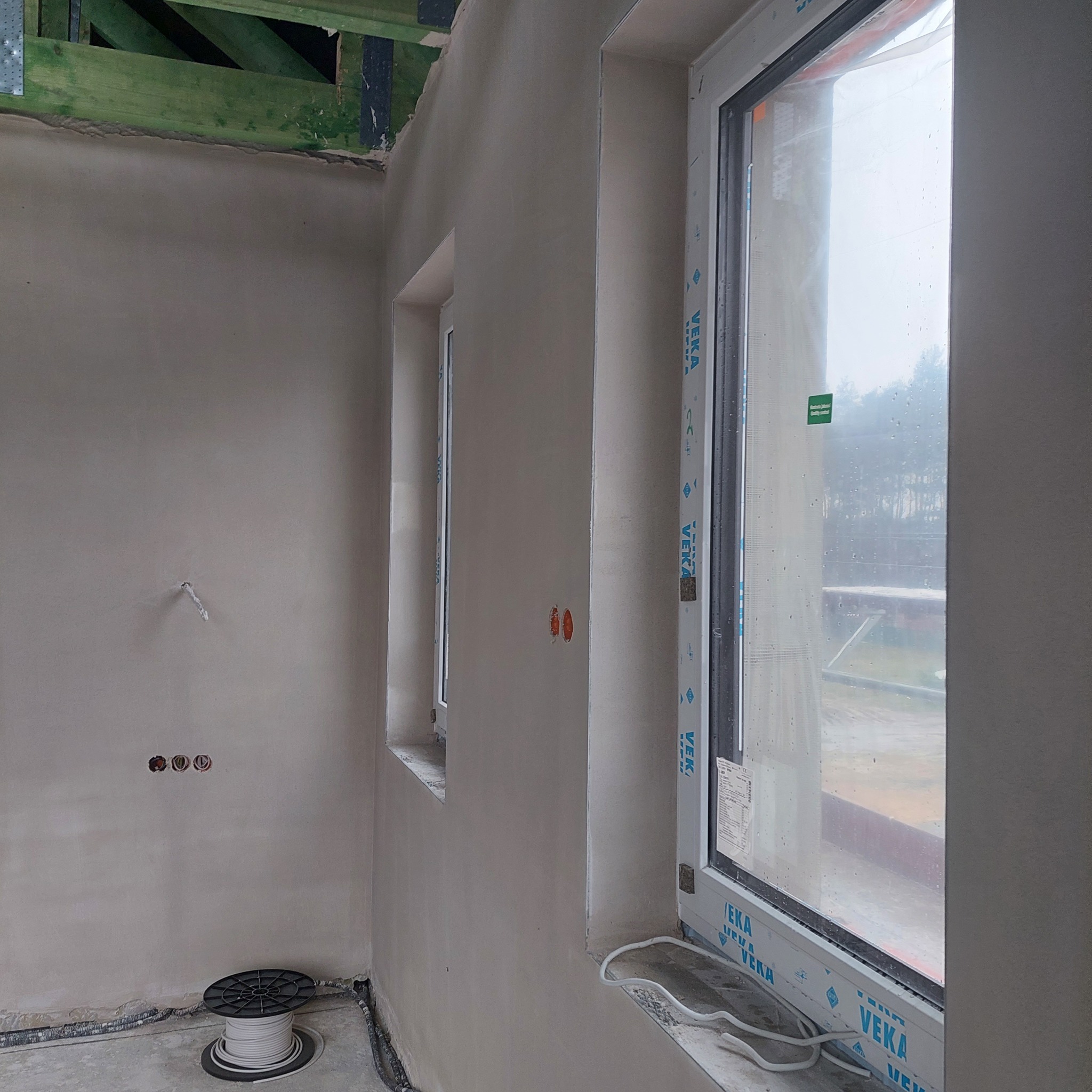 Wnętrze w stanie surowym po nałożeniu świeżych, gładkich tynków na ścianach, widoczne okna PCV z naklejkami producenta, wystające kable elektryczne i puszki instalacyjne oraz rolka kabla na podłodze.