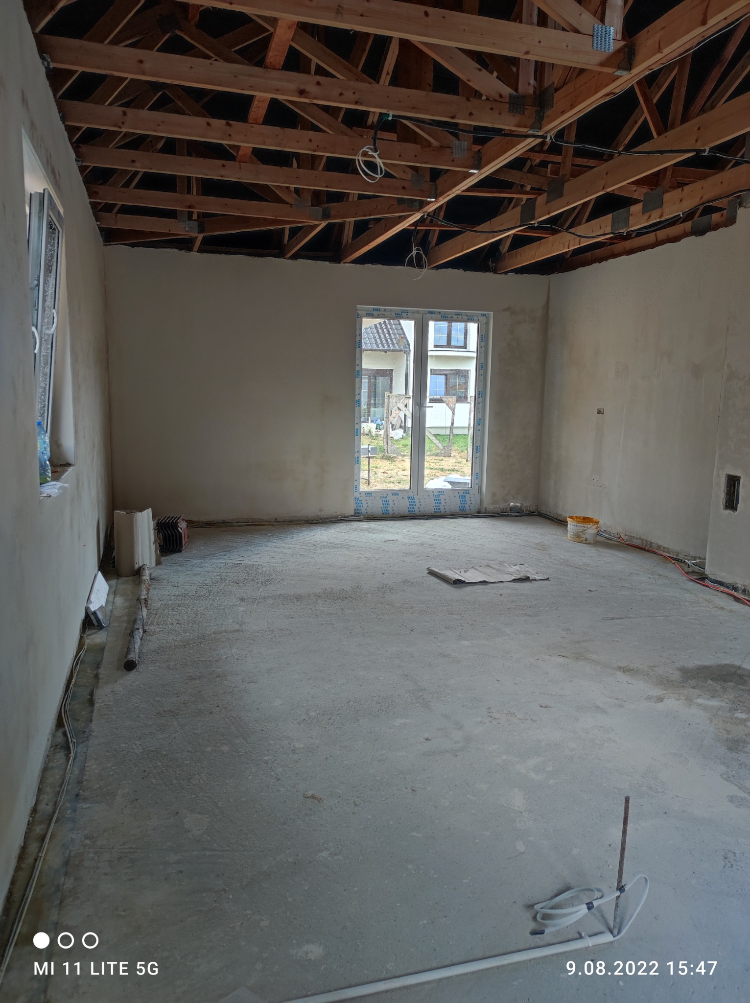 Surowe wnętrze domu w trakcie prac wykończeniowych: ściany pokryte świeżym tynkiem, widoczna drewniana konstrukcja dachu, okno z widokiem na sąsiedni budynek, na podłodze resztki materiałów...