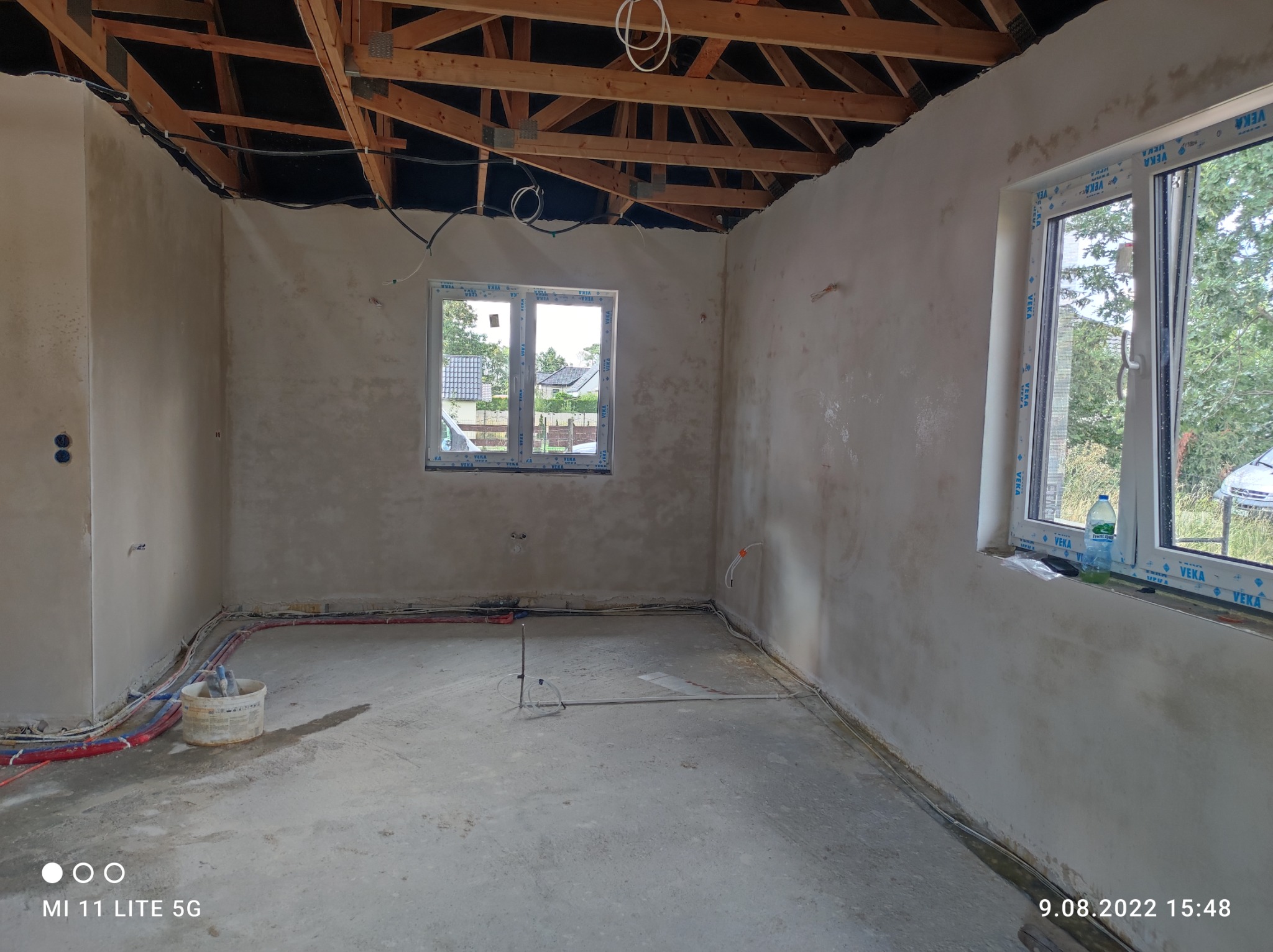 Surowe wnętrze domu w trakcie remontu: ściany po wstępnym tynkowaniu, widoczne instalacje elektryczne i hydrauliczne, surowy strop z drewnianą konstrukcją, okna PCV z widokiem na zieleń.