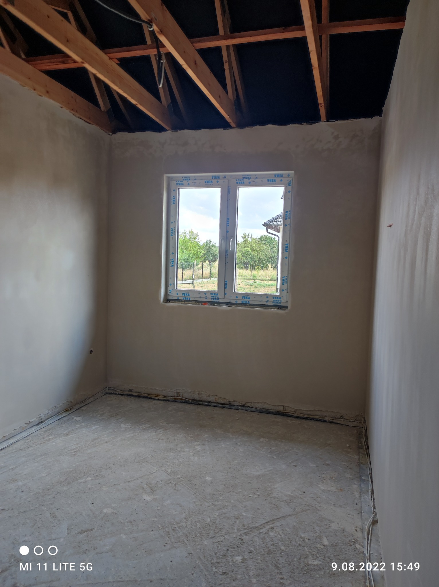 Surowe wnętrze pokoju z oknem, ściany po tynkowaniu, widoczna konstrukcja dachu, podłoga w stanie surowym.