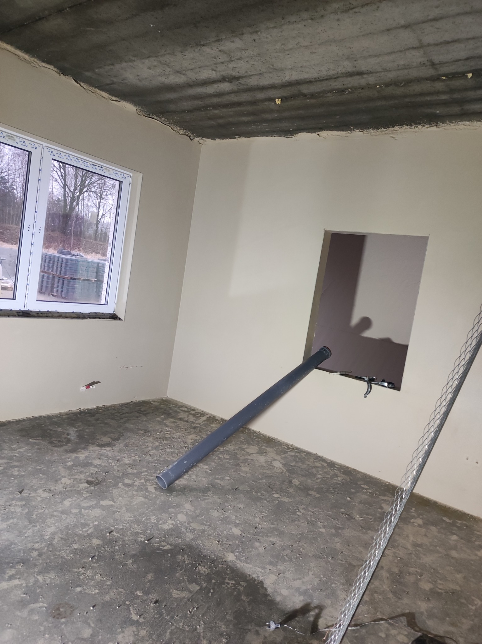 Surowe wnętrze w trakcie prac wykończeniowych: ściany po nałożeniu gładzi, widoczna rura wystająca ze ściany, surowy betonowy sufit i okno z widokiem na drzewa.