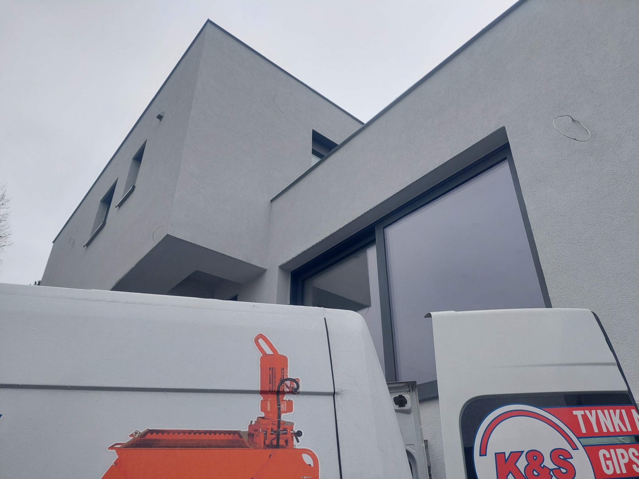 Nowoczesny dom z szarą elewacją, widoczny z perspektywy dolnej, z fragmentem furgonetki z logo firmy 'Tynki Maszynowe K&S Gips' w dolnej części kadru.