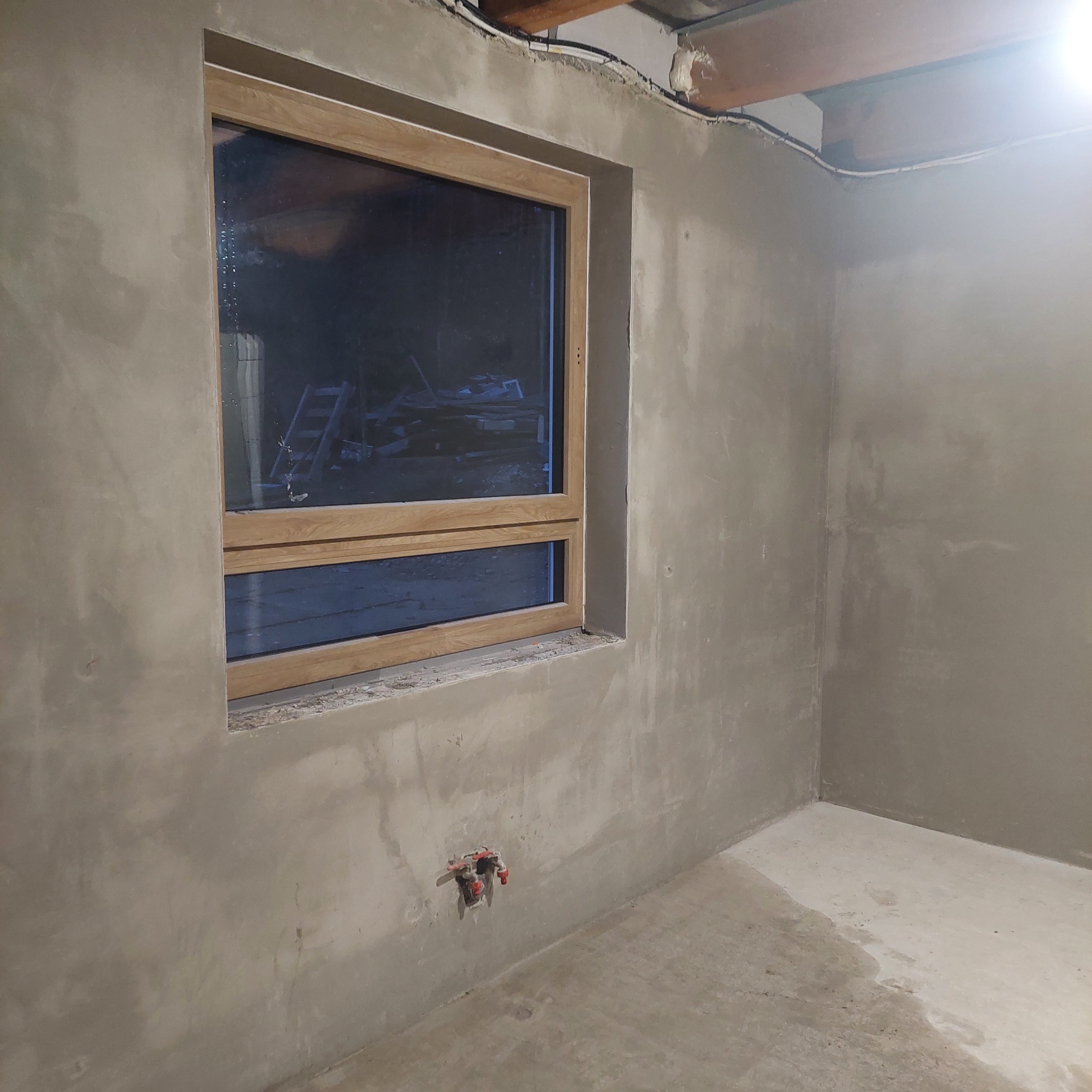 Wnętrze w trakcie prac tynkarskich: ściany pokryte świeżym szarym tynkiem, drewniana rama okienna, widoczne instalacje elektryczne pod sufitem i wystające rury instalacji wodnej.