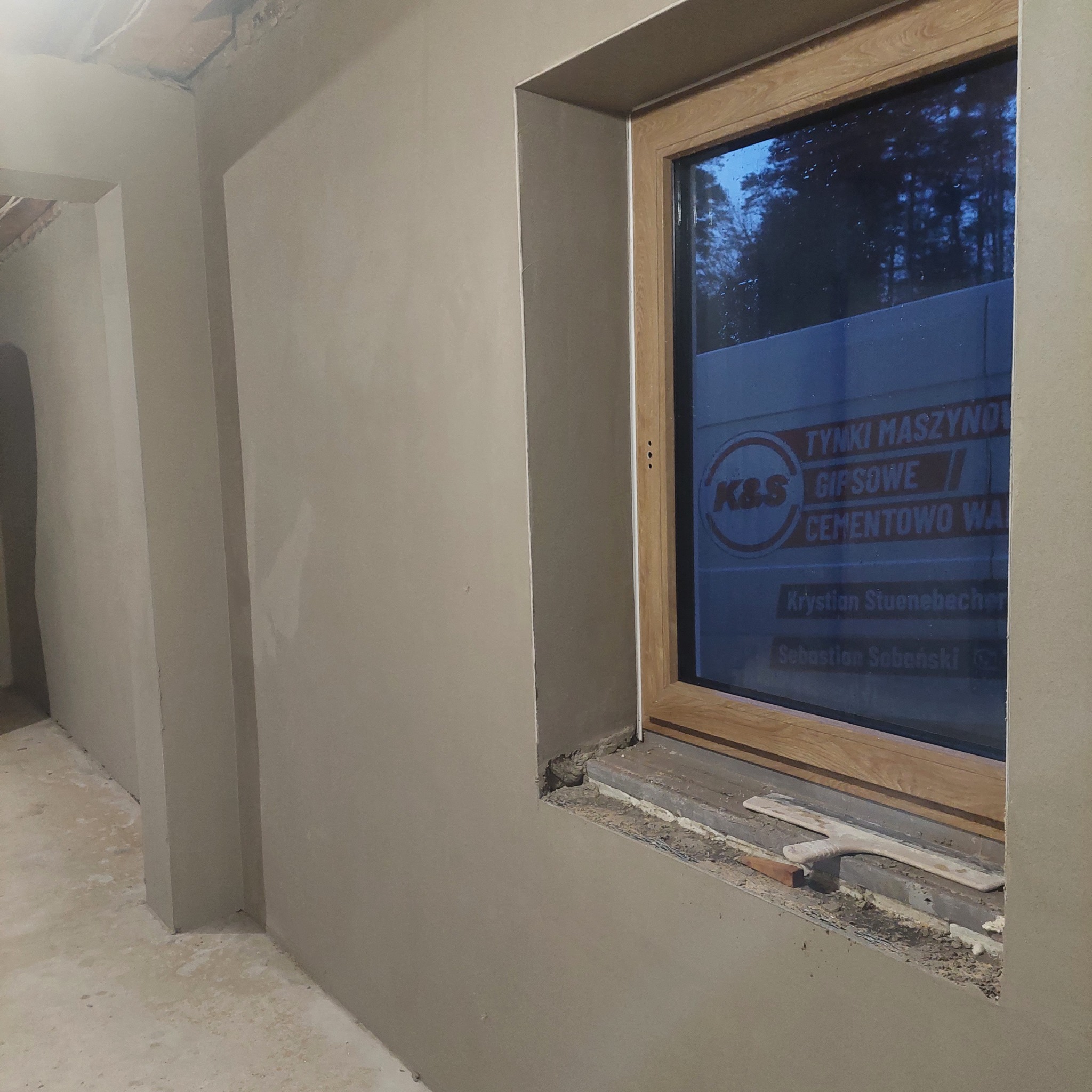 Wnętrze w trakcie prac wykończeniowych: świeżo otynkowane ściany w jasnym odcieniu, okno z drewnianą ramą, na parapecie leży szpachelka, na szybie naklejona reklama firmy budowlanej.