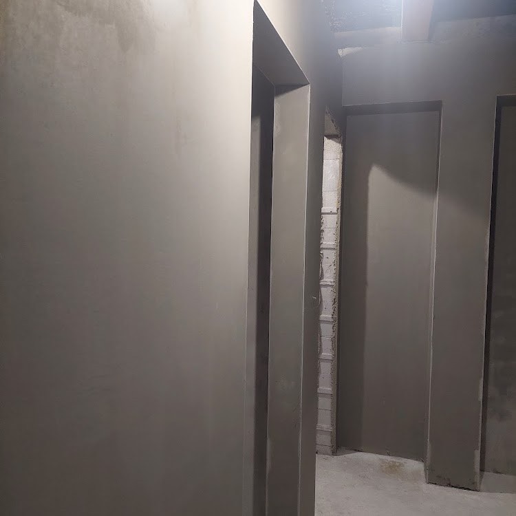 Wnętrze w trakcie prac tynkarskich: świeżo otynkowane ściany w odcieniach szarości, widoczne ościeżnice drzwiowe, surowy mur w prześwicie między pomieszczeniami.