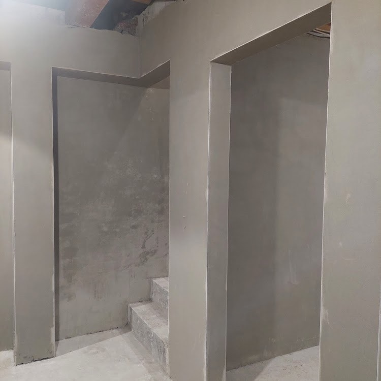 Wnętrze w trakcie prac tynkarskich: ściany i schody pokryte świeżym, jasnoszarym tynkiem, widoczne ościeżnice drzwiowe i surowy strop w tle.