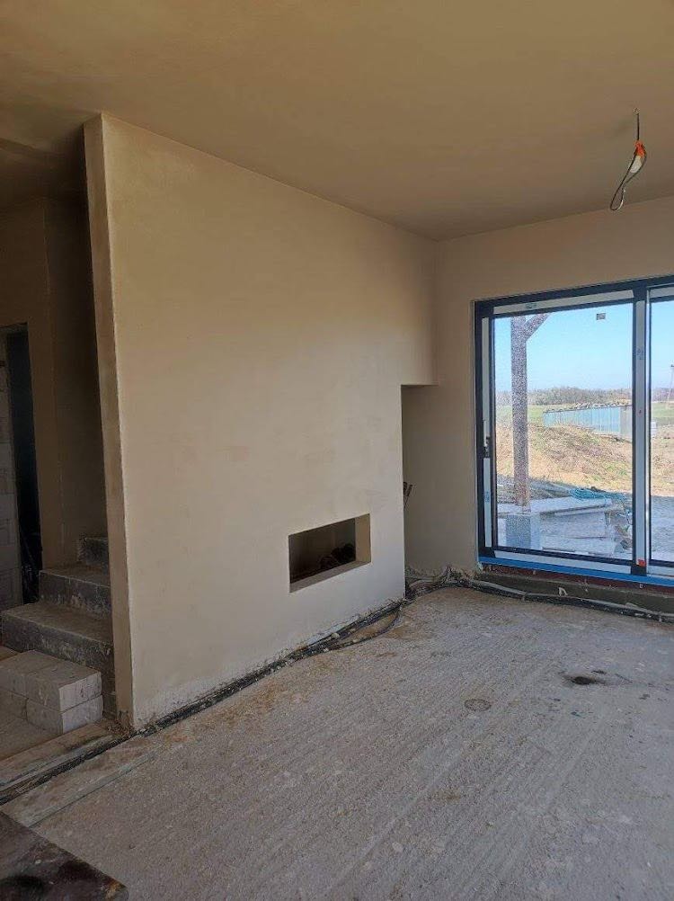 Wnętrze domu w trakcie prac wykończeniowych: ściany pokryte gładkim, jasnym tynkiem, widoczny otwór na kominek, surowa podłoga, okno z widokiem na zewnątrz i wystające kable elektryczne.