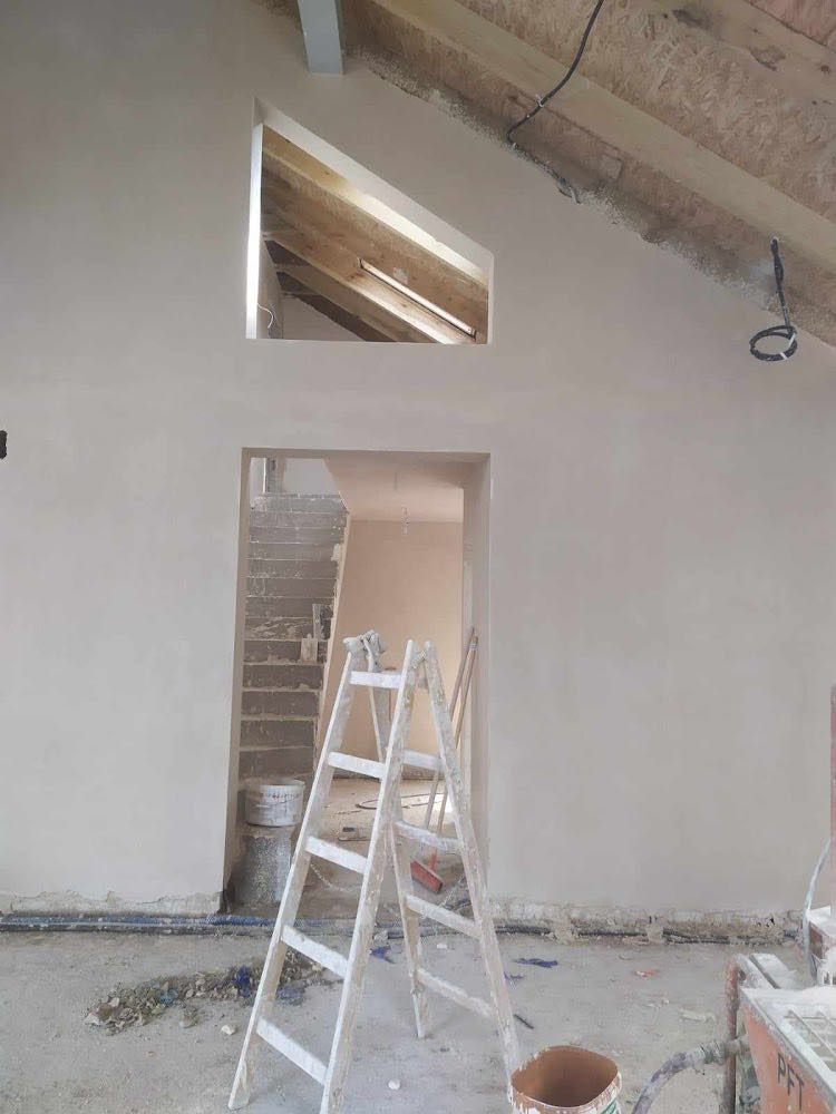 Wnętrze w trakcie prac wykończeniowych: świeżo otynkowana ściana z nietypowym oknem w kształcie trapezu, drewniana konstrukcja dachu widoczna, drabina oparta o ścianę, widoczne ślady po pracach...