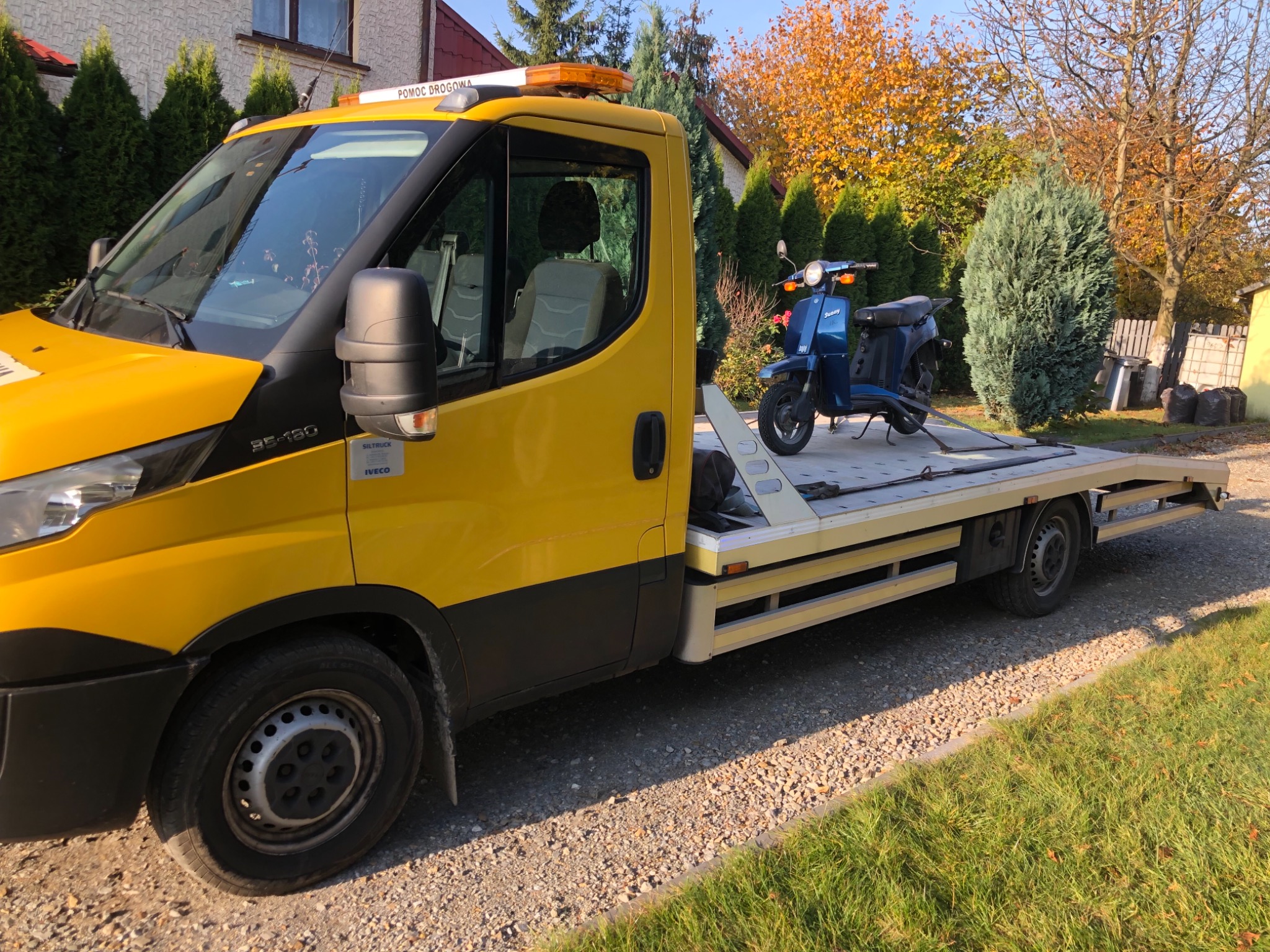 Żółta ciężarówka z platformą przewozi niebieski skuter na tle jesiennego krajobrazu z domem i drzewami.
