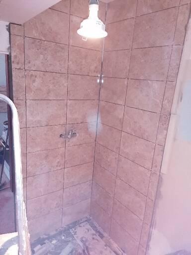 Niedokończona kabina prysznicowa z beżowymi, marmurkowymi płytkami, widoczna instalacja wodna i tymczasowe oświetlenie budowlane.