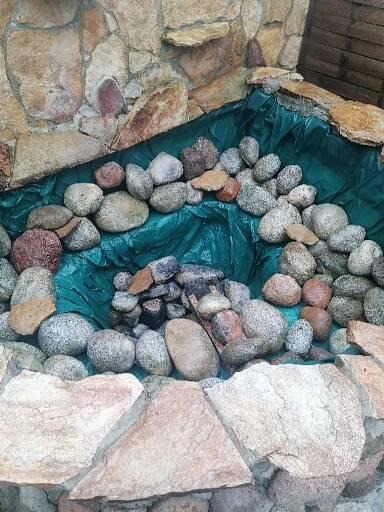 Fragment skalniaka z kamienną ścianą i turkusową folią, wypełnioną różnorodnymi kamieniami w odcieniach szarości, brązu i czerwieni.