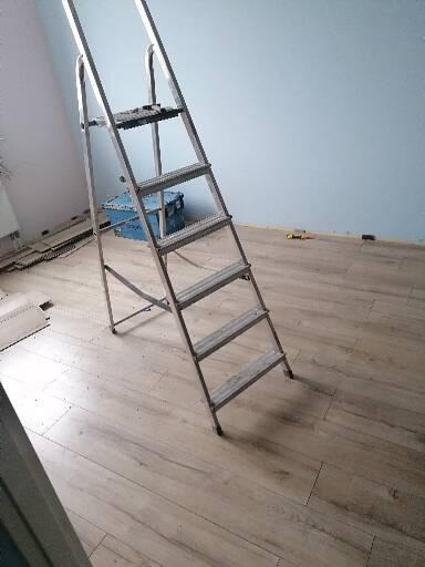 Metalowa drabina stojąca na ułożonych panelach podłogowych, widoczne przygotowane panele do dalszego montażu oraz niebieski pojemnik na narzędzia.