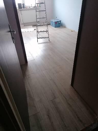 Widok z perspektywy drzwi na świeżo ułożone panele podłogowe w jasnym odcieniu drewna, z drabiną opartą o ścianę i niebieskim pudełkiem w tle, częściowo zakryte folią ochronną.