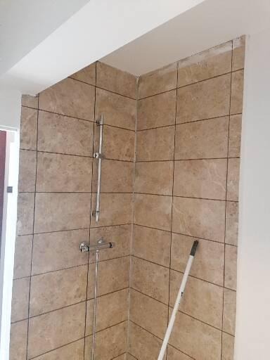 Wykończona kabina prysznicowa z beżowymi, marmurkowymi płytkami, widoczna bateria i drążek prysznicowy oraz biały mop oparty o ścianę.