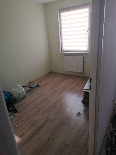 Pokój z ułożoną podłogą z paneli imitujących drewno, widoczne narzędzia i resztki materiałów po pracy, okno z żaluzjami w tle.