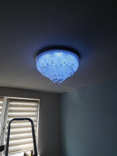 Kryształowy żyrandol sufitowy z niebieskim podświetleniem LED, widoczna drabina malarska przy oknie z roletami.