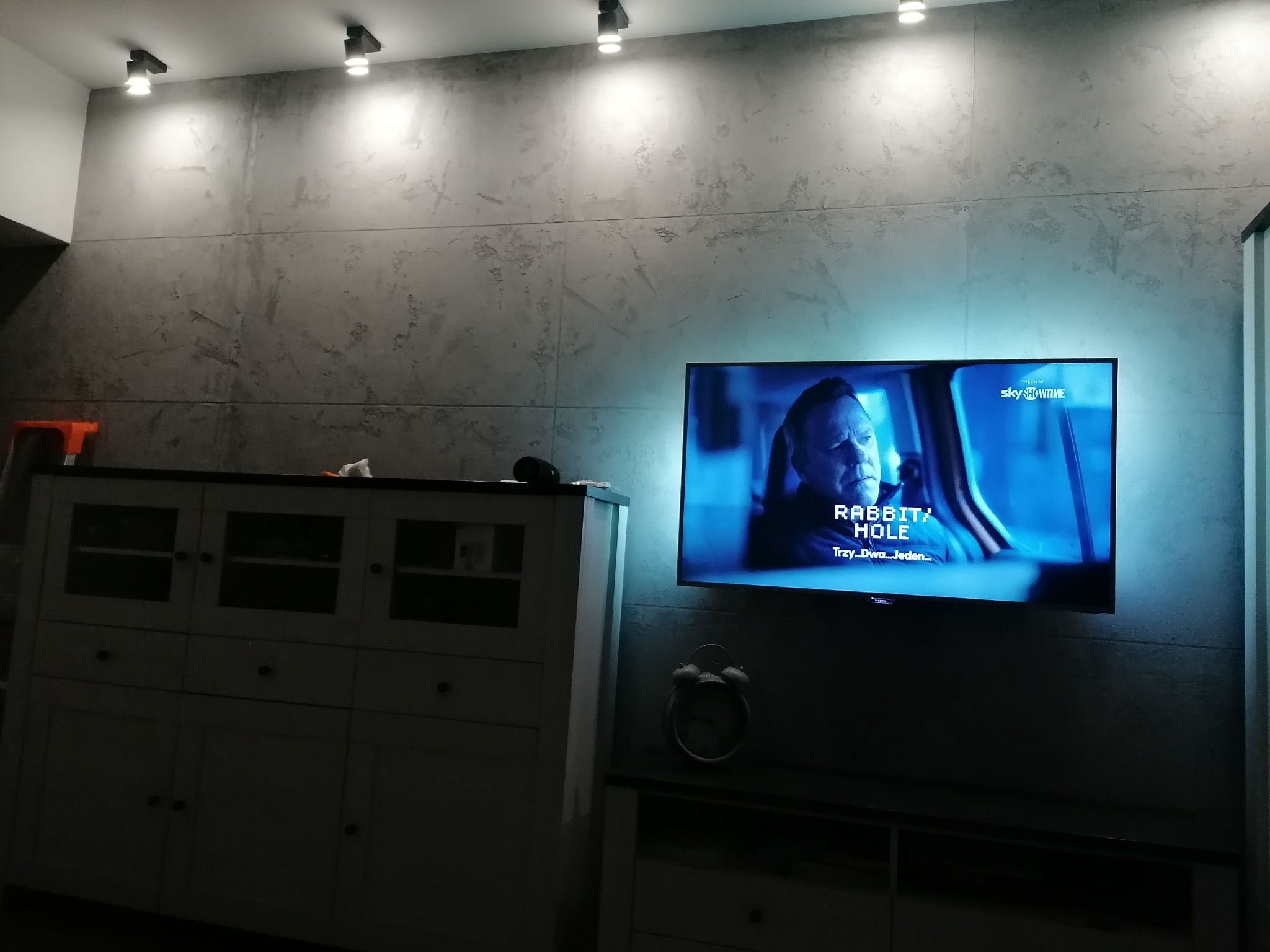 Betonowa ściana z zamontowanymi punktowymi lampami sufitowymi, oświetlona telewizorem z podświetleniem LED, widoczny biały kredens i zegar.