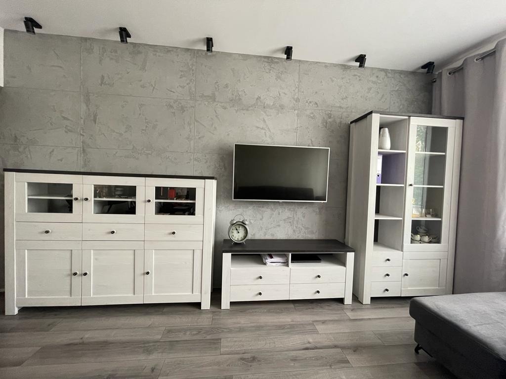 Szara ściana z efektem betonu z zamontowanymi reflektorami sufitowymi, pod nią białe meble z czarnymi blatami: komoda, szafka RTV i witryna, na podłodze panele imitujące drewno.