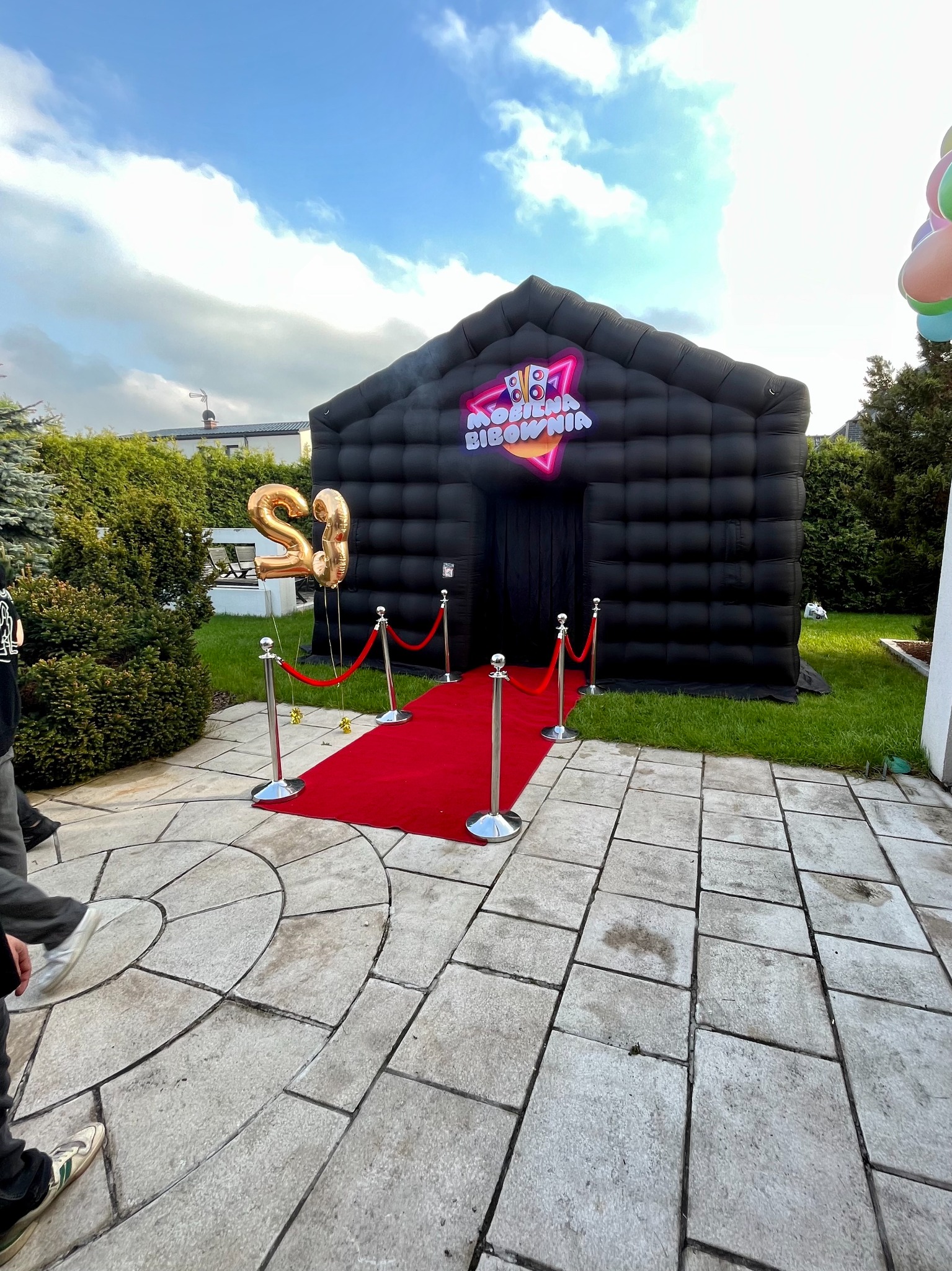 Czarny, dmuchany namiot eventowy z czerwonym dywanem, balonami w kształcie cyfr '23' i napisem 'Mobilna Bibownia' na zewnątrz, ustawiony na trawniku przed budynkiem.