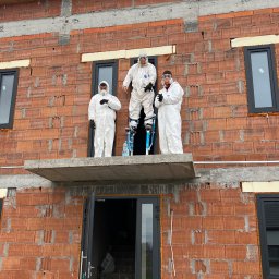 PRO-MAX JAKUB WÓJCIGA - Trzech pracowników w białych kombinezonach ochronnych i maskach, jeden na szczudłach, na tle surowej ściany budynku z czerwonej cegły z widocznymi oknami i betonowymi elementami konstrukcyjnymi.