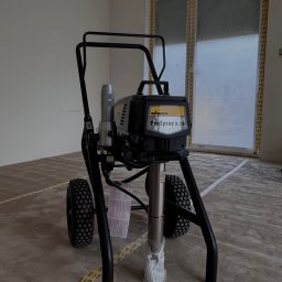 PRO-MAX JAKUB WÓJCIGA - Profesjonalny agregat malarski Wagner ProSpray 3.39 na tle zabezpieczonego folią okna i przygotowanej do malowania podłogi w pomieszczeniu.