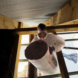 PRO-MAX JAKUB WÓJCIGA - Pracownik w kombinezonie i masce trzyma rolkę wełny mineralnej na poddaszu podczas ocieplania, widoczne drewniane elementy konstrukcyjne i okno.