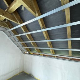 PRO-MAX JAKUB WÓJCIGA - Szkielet pod płytę G-K na drewnianej konstrukcji dachu, widok z dołu. Metalowe profile zamocowane do drewnianych belek. Ściany i podłoga przygotowane do dalszych prac.