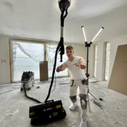 Mężczyzna z szlifierką do gładzi na wysięgniku, w trakcie pracy w jasnym pomieszczeniu. Widoczne lampy, okna zabezpieczone folią i odkurzacz przemysłowy.